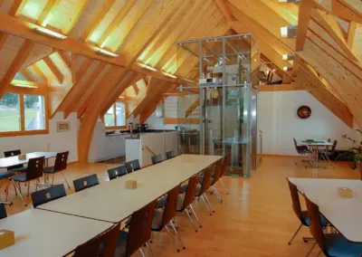 Moderner Konferenzraum im Dachgeschoss mit Holzbalkendecke und Glasaufzug, eingerahmt von Fenstern im Holzrahmen.