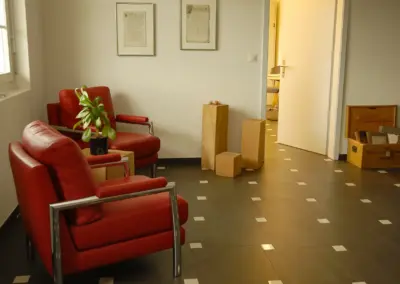 Wartezimmer mit roten Sesseln, Pflanze und moderner Einrichtung, heller Raum mit Kunst an der Wand und offenen Türen.