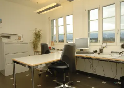 Modernes Büro mit Schreibtischen, Bürostühlen und großem Fenster mit Bergblick. Ideal für produktives Arbeiten.