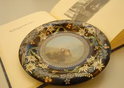 Antiker Teller mit floralem Design auf geöffneter Buchseite, zeigt Landschaft mit Burg und Segelboot.