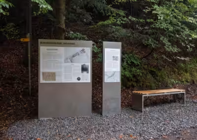 Informationsschild und Bank in der Burgstelle Heimberg, umgeben von Wald. Ideal für Wanderstopps und historische Einblicke.