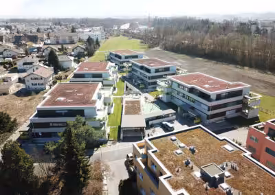 Moderne Wohnanlage mit begrünten Dächern in ländlicher Umgebung, umgeben von Feldern und Bäumen, unter sonnigem Himmel.
