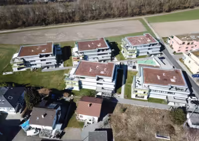Moderne Wohnanlage mit grünen Dächern und Gärten, umgeben von Feldern und Bäumen, aus der Vogelperspektive.