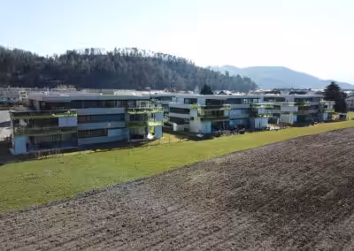 Moderne Wohngebäude neben einem Feld mit Wald im Hintergrund an einem sonnigen Tag.