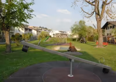 Kinderspielplatz mit Wippe, Schaukeln und Rutsche, umgeben von Bäumen und Wohnhäusern an einem sonnigen Tag.