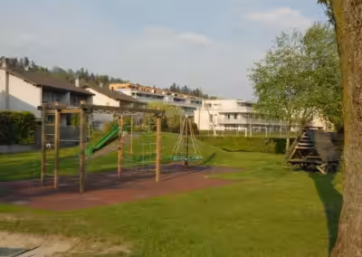 Spielplatz mit Klettergerüst und Rutsche, umgeben von Wohnhäusern und Bäumen an einem sonnigen Tag.