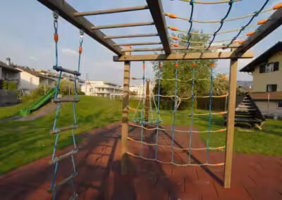 Klettergerüst auf einem Spielplatz mit Seilen und Leitern, umgeben von Rasen, Rutsche und Wohnhäusern im Hintergrund.