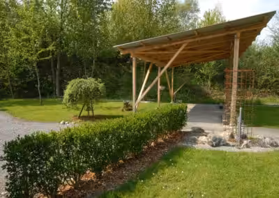 Überdachter Carport aus Holz in einem grünen Garten mit Hecken und Bäumen im Hintergrund bei sonnigem Wetter.