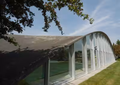 Modernes, geschwungenes Glasgebäude bei sonnigem Himmel, umgeben von grüner Landschaft und Bäumen.