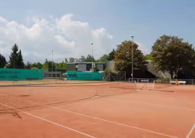 Leerer Tennisplatz bei sonnigem Wetter, umgeben von Bäumen und Bergen im Hintergrund.
