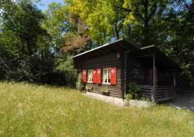 Holzhütte mit roten Fensterläden im Grünen, umgeben von Bäumen und einer Wiese unter blauem Himmel.