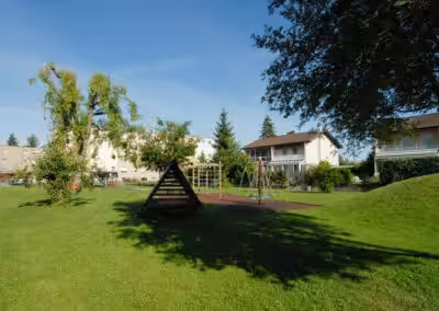 Spielplatz mit Klettergerüst und Schaukeln auf grünem Rasen, umgeben von Wohnhäusern bei sonnigem Wetter.