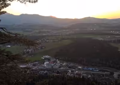 Sonnenuntergang über einem Tal mit Wäldern und Dörfern, gesehen von einem Hügel aus, unter klarem Himmel.