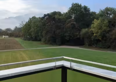 Blick vom Balkon auf grüne Felder und Bäume unter bewölktem Himmel. Natürliche Landschaft in ländlicher Umgebung.