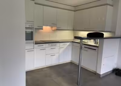 Moderne weiße Einbauküche mit Elektrogeräten und grauem Fliesenboden in einer Wohnung.