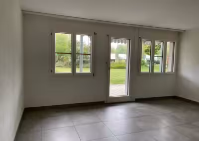 Leerer Raum mit großen Fenstern, die Ausblick auf grünen Garten bieten, moderne Fliesen und viel Tageslicht.