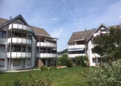 Mehrfamilienhaus mit Balkons, umgeben von grüner Wiese und blühenden Bäumen bei sonnigem Wetter.